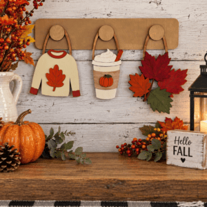 Red Rooster Laser Arts DIY Interchangeable Wall Peg System Decor - Cozy Fall features a charming wooden autumn display with sweaters, pumpkin drinks, leaves, and interchangeable wall hangings. It’s accented by pumpkins, berries, a lantern, candle, Hello Fall sign and other rustic fall elements on a wooden table—perfect for easily updating your decor for patriotic celebrations or any season.