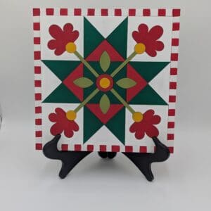 Red Rooster Laser Arts Winter Barn Quilt DIY Craft Kit featuring a square board with a red and green geometric star and flower design, framed by a red and white striped border, displayed on a black plastic stand.