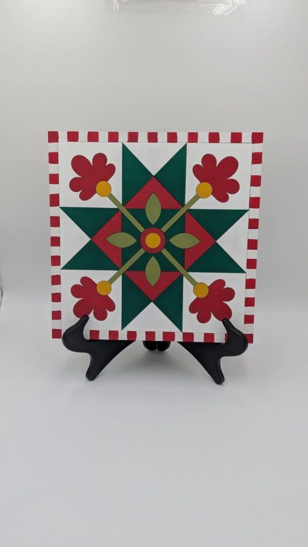Red Rooster Laser Arts Winter Barn Quilt DIY Craft Kit featuring a square board with a red and green geometric star and flower design, framed by a red and white striped border, displayed on a black plastic stand.