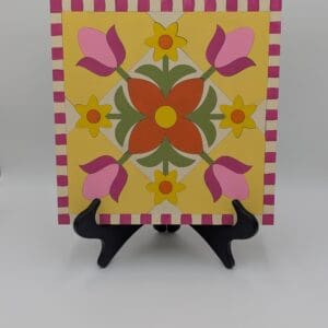 Red Rooster Laser Arts A square wooden panel featuring a painted floral quilt design in yellow, orange, pink, and green, displayed on a black plastic stand as part of a Winter Barn Quilt DIY Craft Kit.