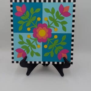 Red Rooster Laser Arts Winter Barn Quilt DIY Craft Kit featuring a floral pattern in shades of blue, pink, and green with a black and white checkered border, displayed on a black stand.