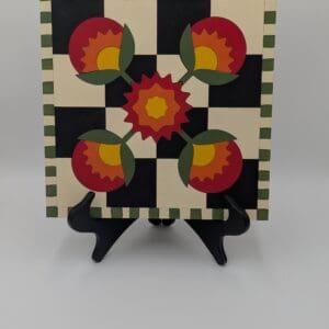 Red Rooster Laser Arts Winter Barn Quilt DIY Craft Kit featuring a square wooden barn quilt with a black and cream checkerboard pattern, a central red and yellow flower motif, and a green striped border, displayed on a black stand.