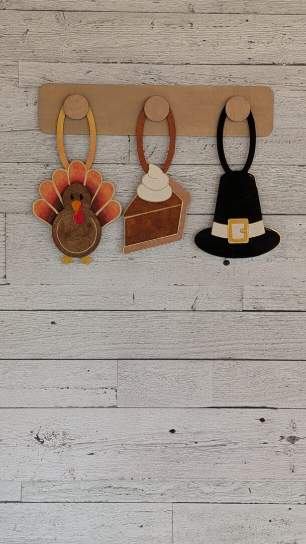 Red Rooster Laser Arts Three wooden ornaments shaped like a turkey, a slice of pie, and a pilgrim hat from the DIY Interchangeable Wall Peg System Decor - Gobble, Gobble and Gather hanging on a wall-mounted peg rack.