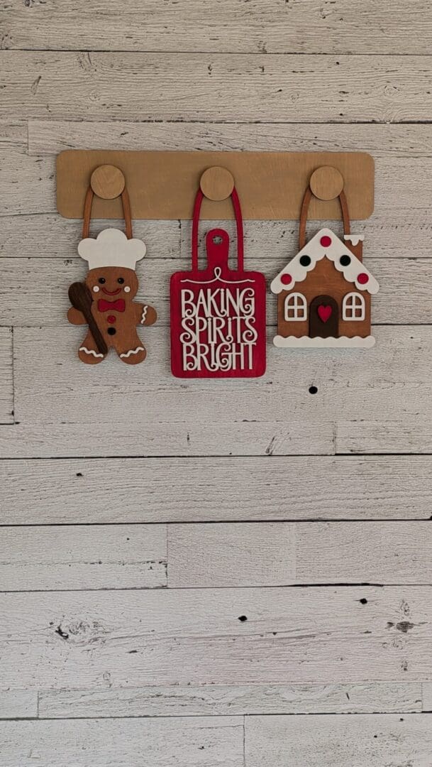 Red Rooster Laser Arts A wooden wall hook rack from the DIY Interchangeable Wall Peg System Decor - Baking Spirits Bright kit holds three holiday ornaments: a gingerbread man with a spoon, a red sign reading Baking Spirits Bright, and a gingerbread house.