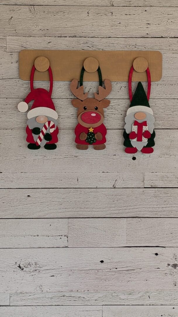 Red Rooster Laser Arts Three Christmas-themed ornaments featuring a gnome with a candy cane, a reindeer, and a gnome with a gift, all hanging from a DIY Interchangeable Wall Peg System Decor - Gnome for the Holidays.